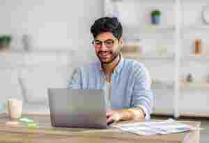 Portrait of happy freelancer man sitting at desk with laptop computer at home office, looking and smiling at screen