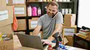 man multitasking on a laptop with a phone at a busy donation center warehouse.