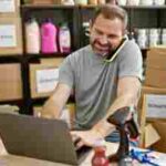 middle-aged man multitasking on a laptop with a phone at a busy donation center warehouse.